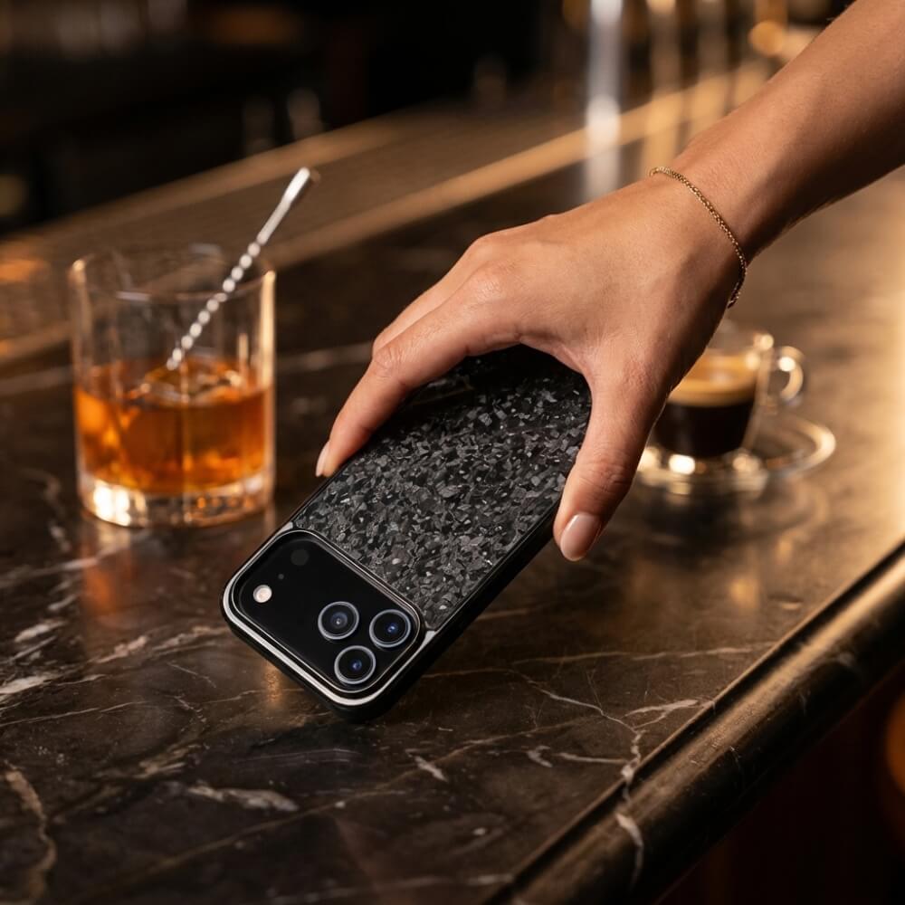A hand with a delicate gold bracelet holds an iPhone featuring a black textured forged carbon case and triple camera array on a dark marble bar next to an amber whiskey with ice and a small espresso