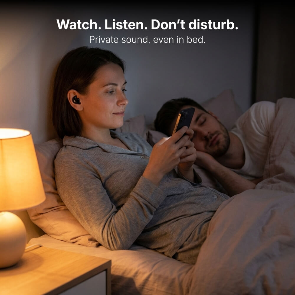 Photograph of a woman in grey pajamas wearing a black wireless earbud, focused on her glowing phone screen while a man sleeps peacefully beside her in a dimly lit bedroom, with a warm bedside lamp