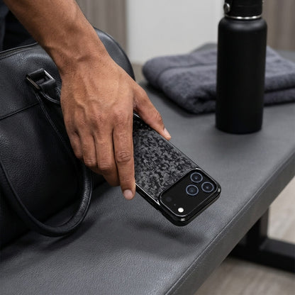 A dark-skinned hand places a smartphone with a forged carbon fiber case and triple camera array onto a gym bench with a black water bottle and grey towel nearby